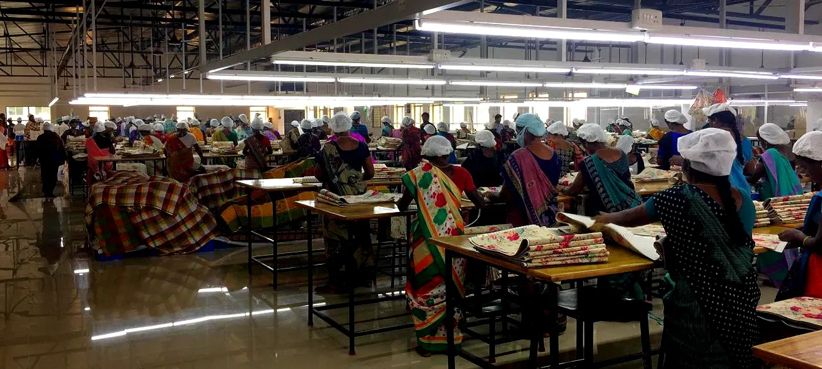 Productiehal in India: rijen werktafels waar medewerkers bedrukte stoffen sorteren en inpakken, onder een open staalconstructie met tl-verlichting.